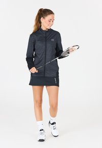 Ung kvinna i svart träningsklädsel som håller en badmintonracket, stående med vita strumpor och skor mot en vit bakgrund.