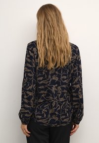 Blouse à manches longues en tissu bleu marine ornée d'un motif floral beige, avec une coupe décontractée et des poignets froncés. Texture lisse avec un léger éclat.