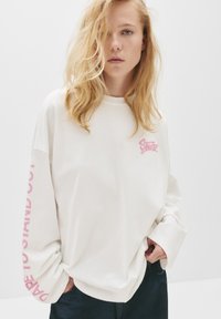 Biała długorękawowa bluza z miękkiego materiału, z różowym logo z przodu i wyboldowanym różowym napisem na lewym rękawie. Luźny krój.