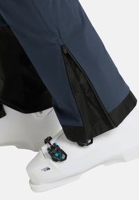 Pantaloni da sci impermeabili color navy con dettagli in cerniera e pannelli neri, dotati di apertura laterale con cerniera sopra un boot da sci bianco con accenti blu.