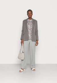 Blazer cinza sobre uma blusa floral, calças largas cinza claro e sandálias creme. A segurando uma bolsa bege clara com uma alça franzida.