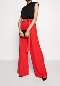 Žena v bezramenném černém topu, širokých červených kalhotách s vysokým pasem a uvázaným opaskem, držící černou psaníčko a černé špičaté boty.