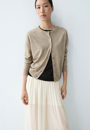 Kobieta ubrana w beżowy, zapinany na guziki sweter na czarnym topie i długą, białą plisowaną spódnicę, stojąca na jednolitym białym tle.