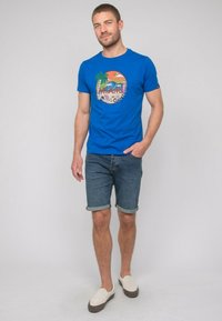 T-shirt bleu royal à manches courtes avec un graphique circulaire de Miami. Associé à un short en denim clair et des chaussures à enfiler crème.