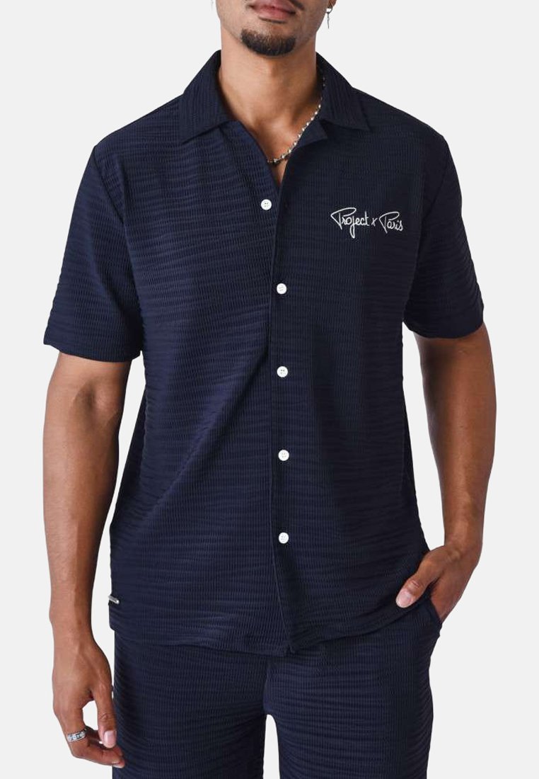 Chemise à manches courtes, à boutons, en tissu texturé bleu marine, avec un logo brodé blanc sur la poitrine et un design de col classique.