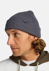 Berretto in maglia grigia con texture a coste, dotato di una piccola etichetta nera sul davanti. Indossato con una felpa con cappuccio marrone.