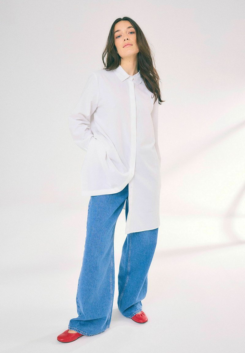 Camicia bianca con colletto e chiusura a bottoni, abbinata a jeans in denim blu a gamba larga e scarpe rosse. Tessuto morbido con una vestibilità rilassata.
