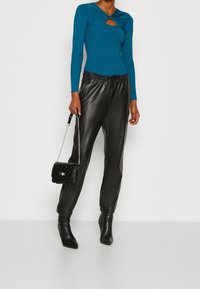 Sinivihreä ribbineulottu pitkähihainen paita, jossa on leikkausdetaili, yhdistettynä mustiin nahkaisiin jogger-housuihin ja mustiin teräväkärkisiin korkokenkiin, kädessä musta tikattu laukku.