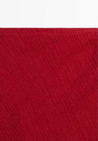 Gros plan sur un tissu rouge texturé avec de subtils plis diagonaux et un bord supérieur cousu sur fond blanc.