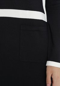 Vêtement en maille noir et blanc avec une bande blanche horizontale et une poche patch, montrant une main près du poignet avec une bordure blanche.