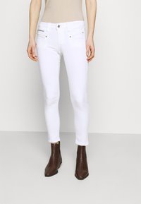 Jeans ajustados blancos con una cintura de media altura, bolsillos con cremallera y acentos de hardware sutil, combinados con botas hasta el tobillo de color marrón oscuro.