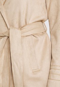 Accappatoio beige realizzato in morbido materiale simile alla pelle scamosciata, con una cintura in vita annodata, tasche laterali e cuciture dettagliate lungo le maniche.