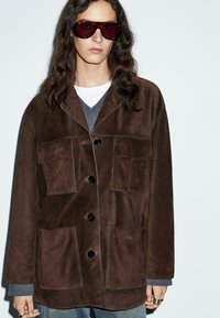 Persona con lunghi capelli scuri che indossa grandi occhiali da sole in tartaruga, giacca scamosciata marrone scuro con grandi tasche, maglione grigio e jeans blu.