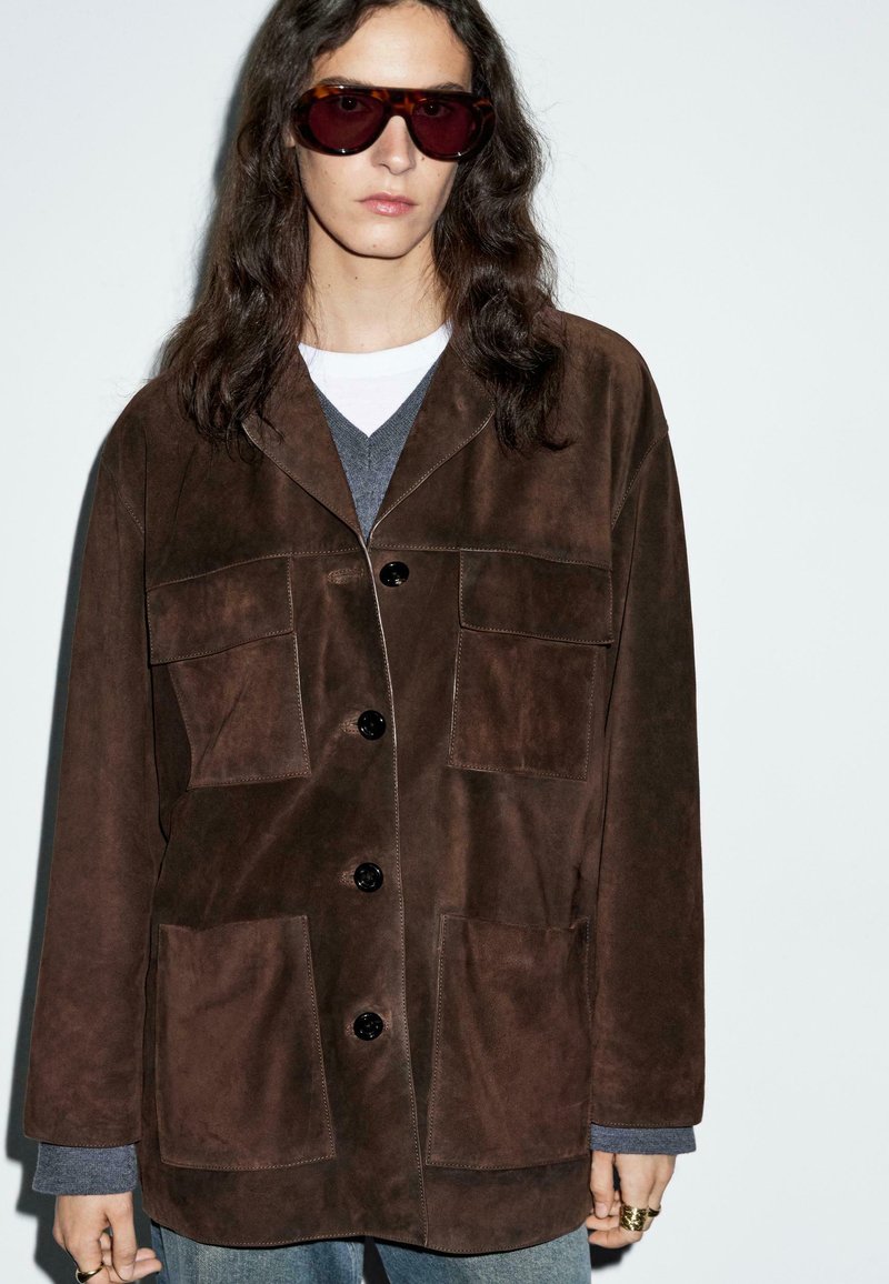 Persona con lunghi capelli scuri che indossa grandi occhiali da sole in tartaruga, giacca scamosciata marrone scuro con grandi tasche, maglione grigio e jeans blu.