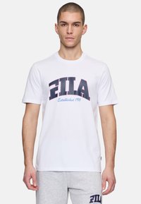 Camiseta de algodón blanca con un audaz logo "FILA" en azul marino y púrpura, con "Establecido en 1911" en una fuente más pequeña debajo. Mangas cortas, cuello redondo.