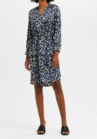 Niebieska i biała sukienka w wzory z długimi rękawami, dekoltem w serek i wcięciem w talii. Noszona z czarnymi sandałami na płaskiej podeszwie. Gładki materiał.