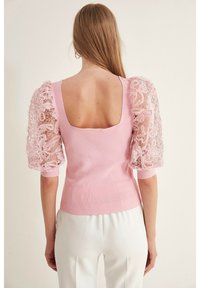 Haut en maille rose avec un dos carré, doté de grandes manches bouffantes en dentelle transparente et d'un ourlet côtelé, offrant un contraste texturé entre les matériaux.