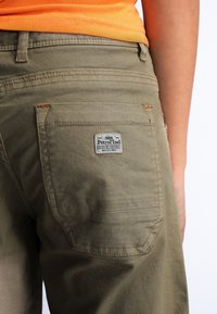 Pantaloni in tela verde con una tasca posteriore che presenta un'etichetta. Il tessuto ha una consistenza liscia e le cuciture sono visibili in filo beige.