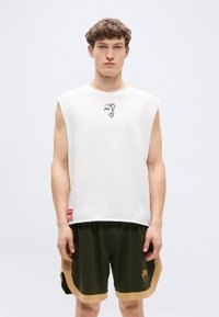 Λευκό αμάνικο μπλουζάκι με γραφικό σχέδιο στο στήθος, συνδυασμένο με σκούρο πράσινο σορτς με ταμπά λεπτομέρεια και λογότυπο κοντά στο τελείωμα.