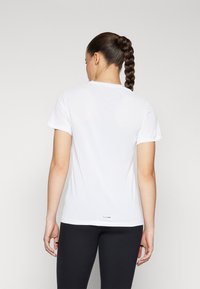 Hvit t-skjorte med kort erm og rund hals, med en glatt tekstur, kombinert med mørke leggins. Logo synlig nær kanten.