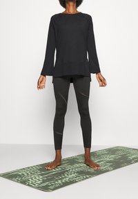 Czarny top z długim rękawem o luźnym kroju, zestawiony z czarnymi legginsami z siateczkowymi detalami. Stojąc na zielonej, wzorzystej macie do jogi.