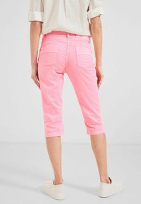 Mujer de pie con pantalones rosas de longitud media, camisa blanca arremangada y zapatillas blancas contra un fondo liso.