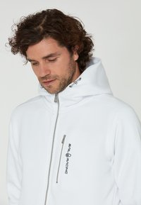 Bílá kapucňová bunda vyrobená z měkké látky, s předním zipem, bočními kapsami a logem "SAIL RACING" na hrudi.