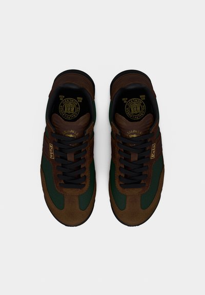 Tenisky s hnědým semišovým svrškem, zelenými textilními doplňky, černými tkaničkami a zlatým logem na jazyku. Plochá gumová podrážka.