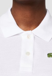 Valkoinen polo-paita, jossa on teksturoitu kangas, klassinen kaulus, kolmen napin mekaaninen sulkeminen ja pieni vihreä brodeerattu krokotiililogot.