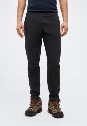 Férfi fekete nadrágot és barna túracipőt visel, és egy tiszta fehér háttér előtt áll.