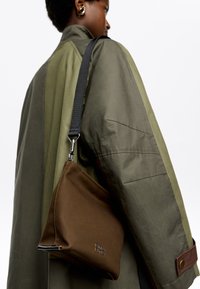 Borsa a mano in pelle marrone con forma triangolare e tracolla nera, dotata di accessori metallici e logo a rilievo, indossata con un cappotto verde oliva.