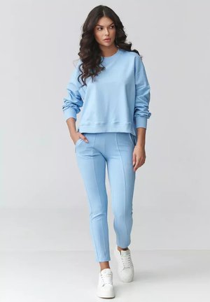 Kobieta o długich, ciemnych, kręconych włosach, ubrana w jasnoniebieską bluzę i dopasowane spodnie, stojąca z jedną ręką w kieszeni, w białych trampkach.