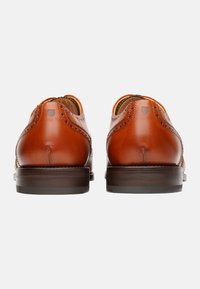 Scarpe di pelle marroni con una finitura liscia, dettagli brogue, tacco arrotondato e una suola di un marrone scuro a contrasto. La vista posteriore mette in mostra le cuciture e gli accenti.