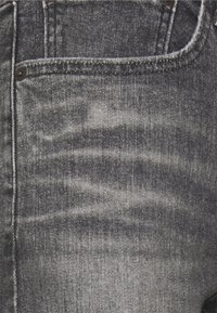 Jeans de denim cinza com um padrão levemente desbotado, apresentando um bolso frontal, costura contrastante e detalhes em botão metálico.