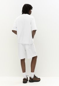 Haut blanc à manches courtes avec une coupe décontractée, associé à un short assorti. Poche latérale visible, porté avec des chaussures de sport marron et des chaussettes blanches.