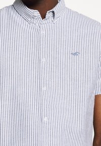 Košile na knoflíky s krátkým rukávem, která má modro-bílé vertikální pruhy, límec s knoflíky a malé vyšívané logo na hrudi.