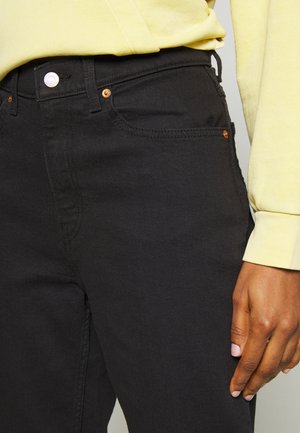 Jeans en denim noir avec une coupe slim, featuring des poches surpiquées, un bouton argenté et des rivets en cuivre. Associés à un haut à manches longues jaune.