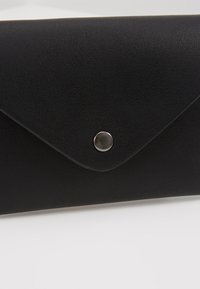 Portefeuille noir texturé de style enveloppe avec une fermeture par bouton-pression argenté sur une surface blanche.