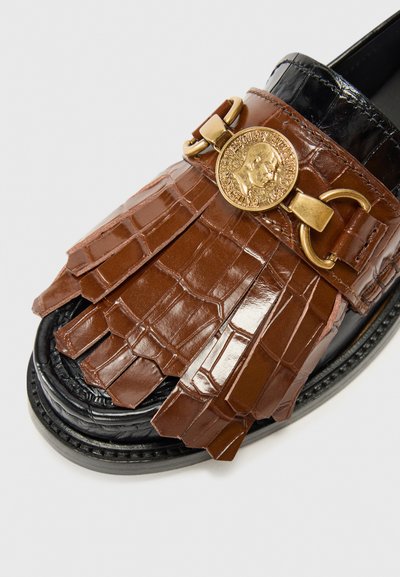 Brown loafers with a textured croc-patterned upper, gold embellishment, and fringe detail, resting on a black sole against a white background.