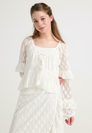 Blusa bianca in pizzo con design a peplum, maniche lunghe volant e scollatura a V. Presenta una chiusura con fiocco e un motivo floreale su tutta la superficie.