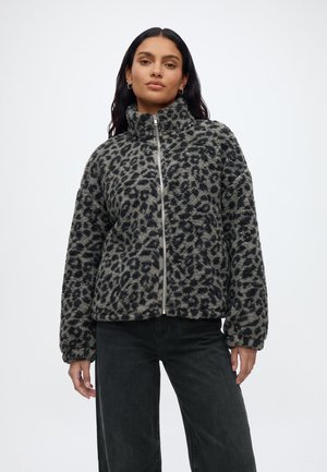 Kvinde iført en grå og sort leopardmønstret fleecejakke med lynlås og lange ærmer samt sorte bukser, stående foran en ensfarvet baggrund.