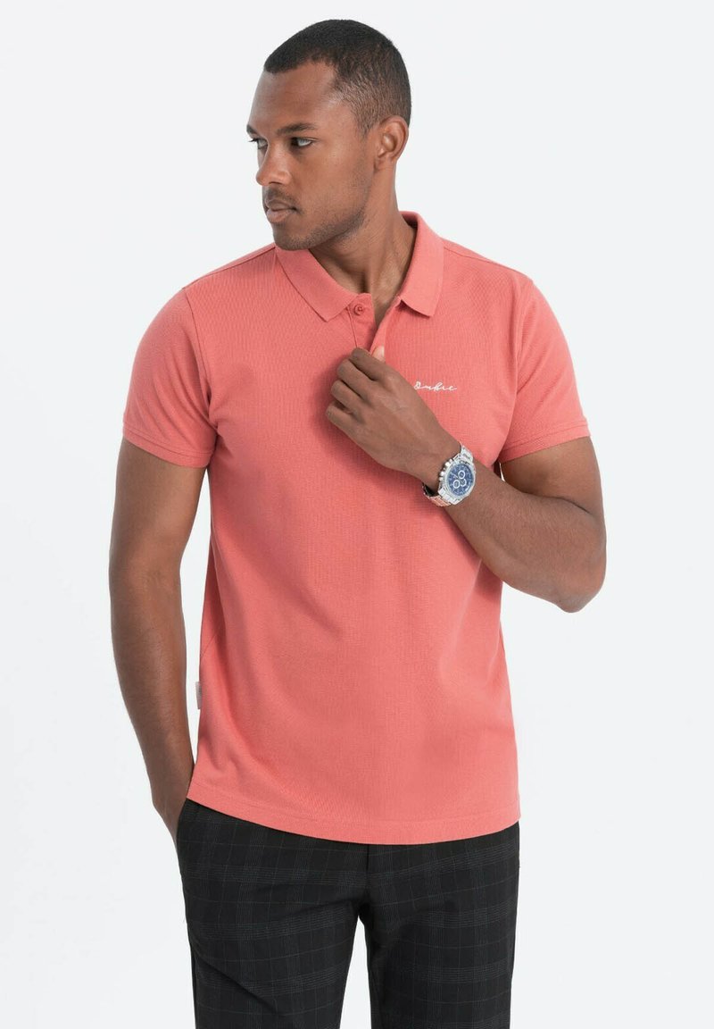 Korallinvärinen polo-paita, jossa on teksturoitu pinta, lyhyet hihat ja kaulus. Sisältää huomaamattoman valkoisen logon rinnassa. Yhdistetty ruudullisiin mustiin housuihin.