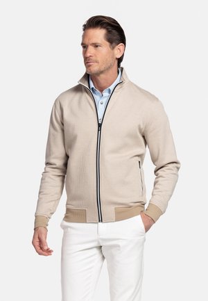 Man wearing beige zip-up jacket over light blue shirt and white pants, standing with one hand in pocket, looking to the side.