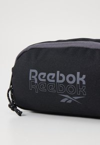 Fekete Reebok tasak, textúrált szövetből készült, szürke logónyomattal. Húzózsinórral záródik, és kontrasztos szürke díszítéssel rendelkezik.