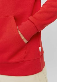 Sudadera roja con un bolsillo tipo canguro, puños ribeteados y dobladillo. La tela parece suave con una textura lisa. Etiqueta lateral visible.