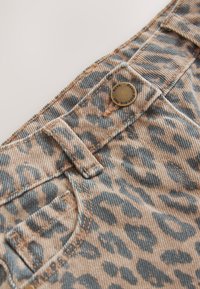 Jeans tkanina z vzorcem leopardjega tiska, prikazana je bližnja slika žepov, šivov in kovinskega gumba z vgravirano podrobnostjo logotipa. Barve vključujejo rjavo in sivo.