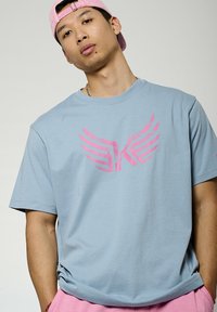 Lichtblauw katoenen t-shirt met een groot roze vleugelt ontwerp op de borst. Losse pasvorm met korte mouwen.