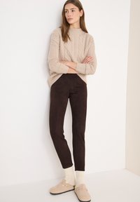 Beżowy sweter w warkocze z wysokim kołnierzem, zestawiony z ciemnobrązowymi legginsami zamszowymi i jasnymi, dzianinowymi skarpetami, uzupełniony beżowymi klapkami.