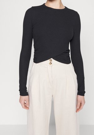 Mujer con un top negro ajustado de manga larga con dobladillo retorcido y pantalones blancos sueltos de talle alto con dos botones delanteros.