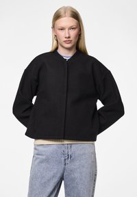 Veste bomber en laine noire avec un col rond, des épaules tombantes et des poches latérales, portée par-dessus un léger pull en maille et un jean en denim.